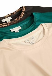 Kolme lyhythihaista puseroa pinottuna: musta leopardi-kuvioinen, teal-värinen ja beige. Suora kangas, pyöreät pääntiet, tarrat näkyvissä kaikissa.