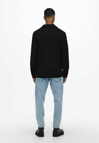 Camisa negra de manga larga con cuello, combinada con unos vaqueros de corte relajado en azul claro y botas negras. Diseño minimalista, sin patrones visibles.