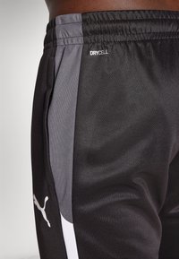 Crne sportske hlače s sivim bočnim panelima i bijelim akcentima. Sadrži "DRYCELL" logo, elastičnu struk i glatku, laganu tkaninu.