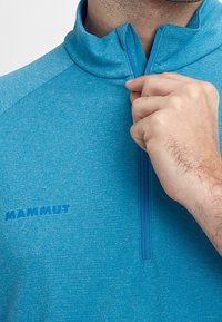 Ljusblå zip-jacka i en strukturerad tyg, med hög krage och "MAMMUT" logotyp tryckt i mörkblått på framsidan.