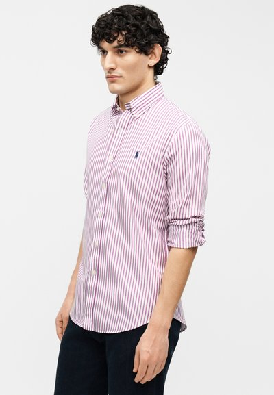 Άνδρας με σγουρά μαλλιά, που φοράει λευκό πουκάμισο με μοβ ριγέ κουμπιά και σκούρο παντελόνι, στέκεται με τα χέρια χαλαρά σε απλό φόντο.