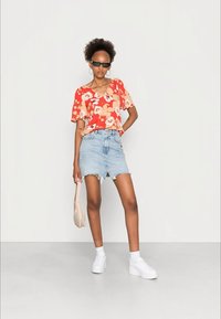 Blusa floral vermelha com mangas curtas, combinada com uma mini saia de ganga desgastada. A modelo usa ténis brancos e segura uma pequena mala de creme.