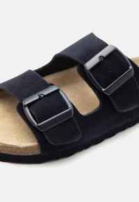 Μαύρο σουέτ σανδάλι με δύο ρυθμιζόμενες μεταλλικές αγκράφες και ταμπά μαλακό πέλμα σε μαύρη σόλα.