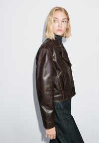 Braune Lederjacke mit kurzem Schnitt, ausgestaltet mit einem Reverskragen, Vorderseitentaschen und glatter Textur. Model trägt graues Oberteil und Jeans.