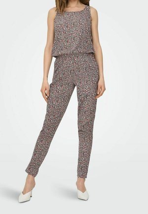 Kobieta w bezrękawniku w kwiatowy kombinezon z nogawkami zwężanymi ku dołowi oraz białe buty na szpic na tle jednolitego tła.