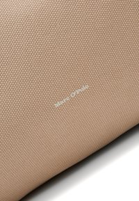 Gros plan sur un tissu beige texturé avec un petit motif tissé uniformément et le texte blanc "Marc O’Polo" centré en diagonale.