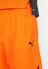 Short de sport orange vif avec logo Puma noir et poche latérale, porté avec un haut noir affichant le texte "DRYCELL" sur la manche.
