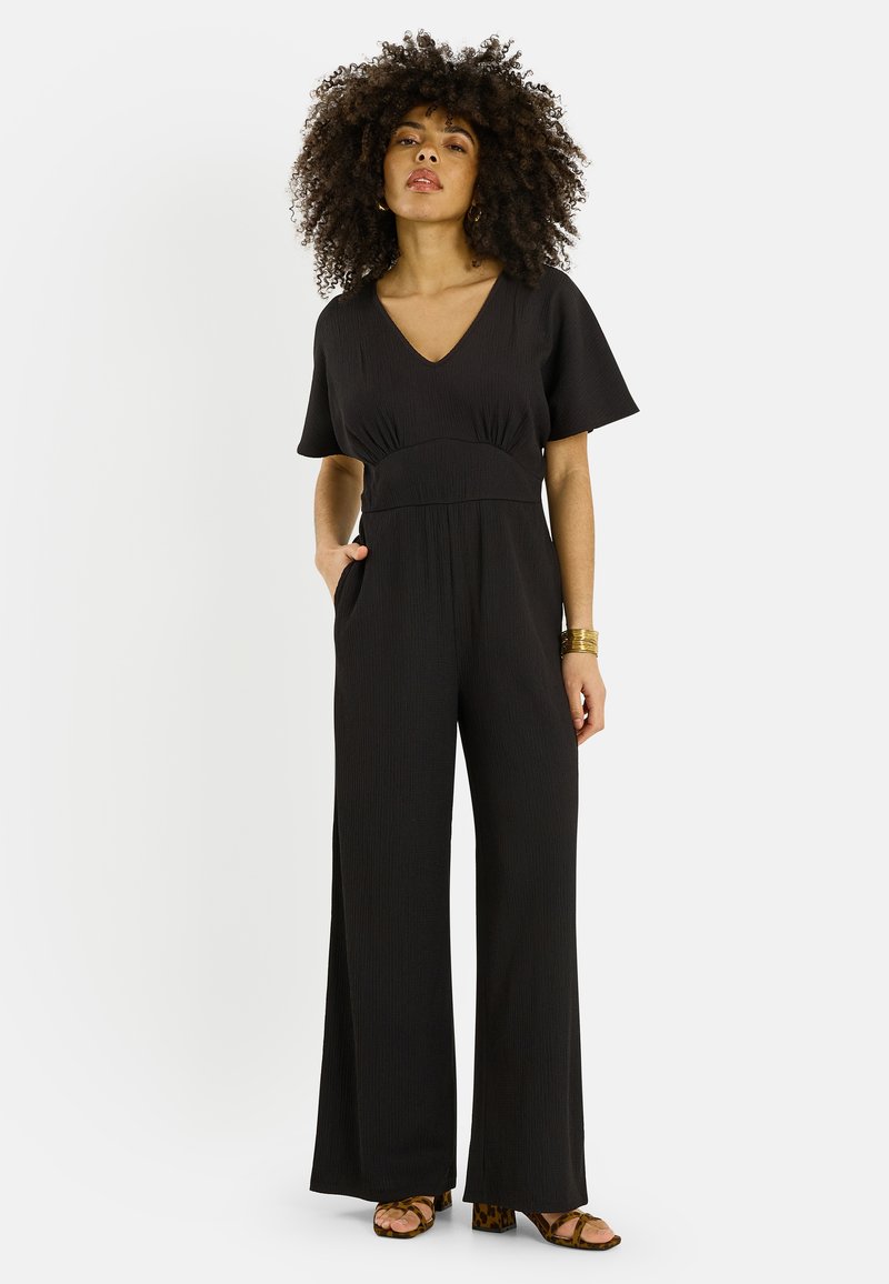 Femme aux cheveux bouclés portant une combinaison noire à manches courtes et jambes larges avec des poches, ainsi que des sandales à talons marron, debout devant un fond blanc.