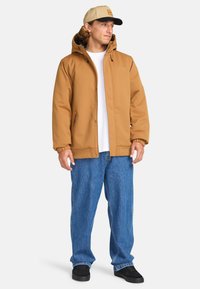 Giacca cappuccio marrone, isolata, con zip e tasche laterali, indossata sopra una t-shirt bianca, abbinata a jeans blu larghi e scarpe nere.