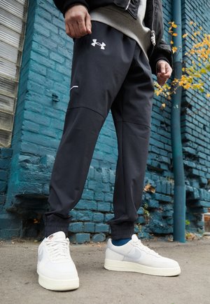 Man in zwarte Under Armour joggers en witte Nike sneakers staat tegen een blauwe bakstenen muur met herfstbladeren in de buurt.
