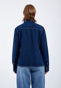 Dunkelblaue Jeanshemd mit langen Ärmeln, Knopfmanschetten und klassischem Kragen. Glatte Textur mit sichtbaren Nähten und einem leicht ausgestellten Saum.