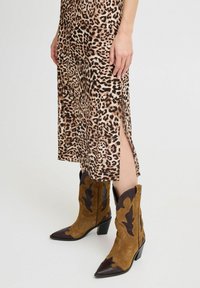 Stivali cowboy in suede marrone e marrone scuro, con punta appuntita, dettagli scallop e tacco spesso, abbinati a un vestito con stampa leopardata.