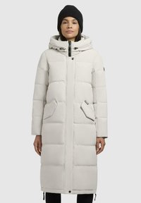 Polstret lang frakk i off-white med hette med snøring, sidelommer, frontlås med glidelås, og svarte detaljer. Strikket svart lue på modellen.