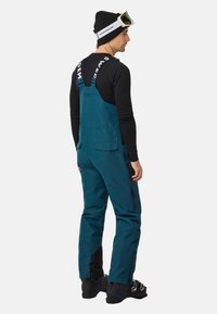 Teal skikleding met verstelbare banden, waterdicht materiaal, zijzakken met rits en zwarte accenten. Gemaakt in combinatie met een zwart shirt met lange mouwen en een beanie.