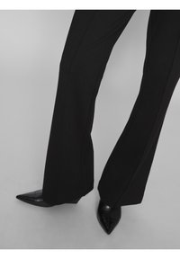 Svarta skräddarsydda byxor med en utsvängd form och vertikala sömmar, tillsammans med spetsiga svarta klackar. Slät yta och enhetlig färg.