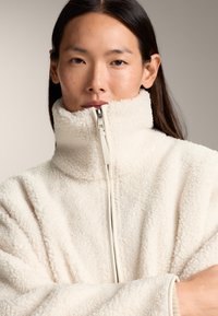 Donna con lunghi capelli scuri che indossa una giacca di pile color crema con collo alto chiusa fino al mento, rivolta in avanti con le braccia incrociate.