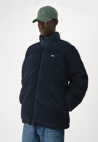 Marine blå fleecejakke med høy krage, glidelås og sidelommer. Har en liten tri-farge logo på brystet og et teksturert stoff.