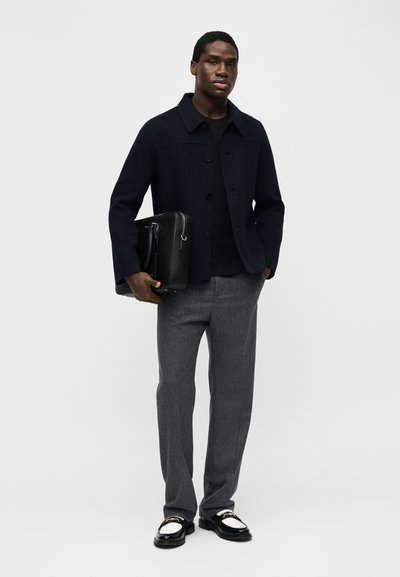 Άνδρας που φοράει ναυτικό μπουφάν, μαύρο πουκάμισο, γκρι παντελόνια, κρατώντας μαύρη δερμάτινη τσάντα, στέκεται μπροστά σε ανοιχτό γκρι φόντο.