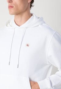 Vyras dėvi baltą gobtuvinį džemperį su „Calvin Klein“ logotipo emblema ant krūtinės, ranka kišenėje priekyje, veidu į kairę, priešais vientisą foną.