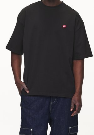 Persoon draagt een zwarte oversized T-shirt met een klein rood-wit "P"-logo op de borst en donkerblauwe cargobroek, hand in de zak.