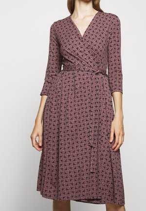 Femme portant une robe portefeuille longueur genou à manches longues avec un motif tourbillonnant rose et foncé, attachée à la taille avec une ceinture assortie.