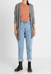 Szary kardigan z dzianiny na pomarańczowo-brązowej koszuli w paski, do tego jasnoniebieskie, podwinięte dżinsy i czarne, sznurowane botki.