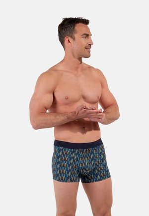 Oberkörperfreier Mann steht, trägt blaue gemusterte Boxershorts, blickt nach rechts, die Hände vor der Taille verschränkt, vor einfachem Hintergrund.