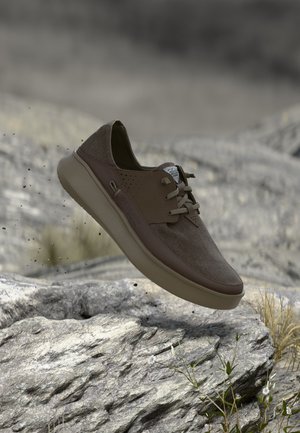 Ανοιχτό καφέ καθημερινό παπούτσι με κορδόνια που αιωρείται πάνω από πετρώδες έδαφος με μικρά φυτά και θολό φόντο.