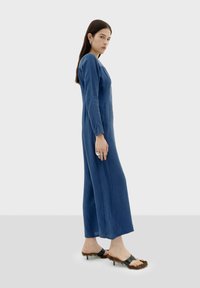 Langsleeve, diepblauwe jurk met een rechte silhoutte, gemaakt van zachte stof. Gecombineerd met zwarte sandalen met gestructureerde accenten.