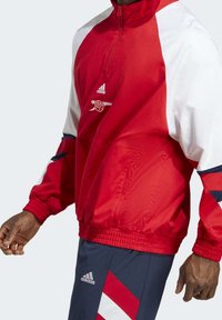 Chaqueta Adidas roja y blanca con media cremallera, puños y dobladillo elásticos. Presenta un logo en el pecho y un diseño en bloques de color con acentos en azul marino.