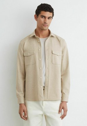 Uomo con capelli corti e ricci che indossa una camicia beige abbottonata sopra una maglietta bianca e pantaloni bianchi, in piedi davanti a una parete bianca semplice.