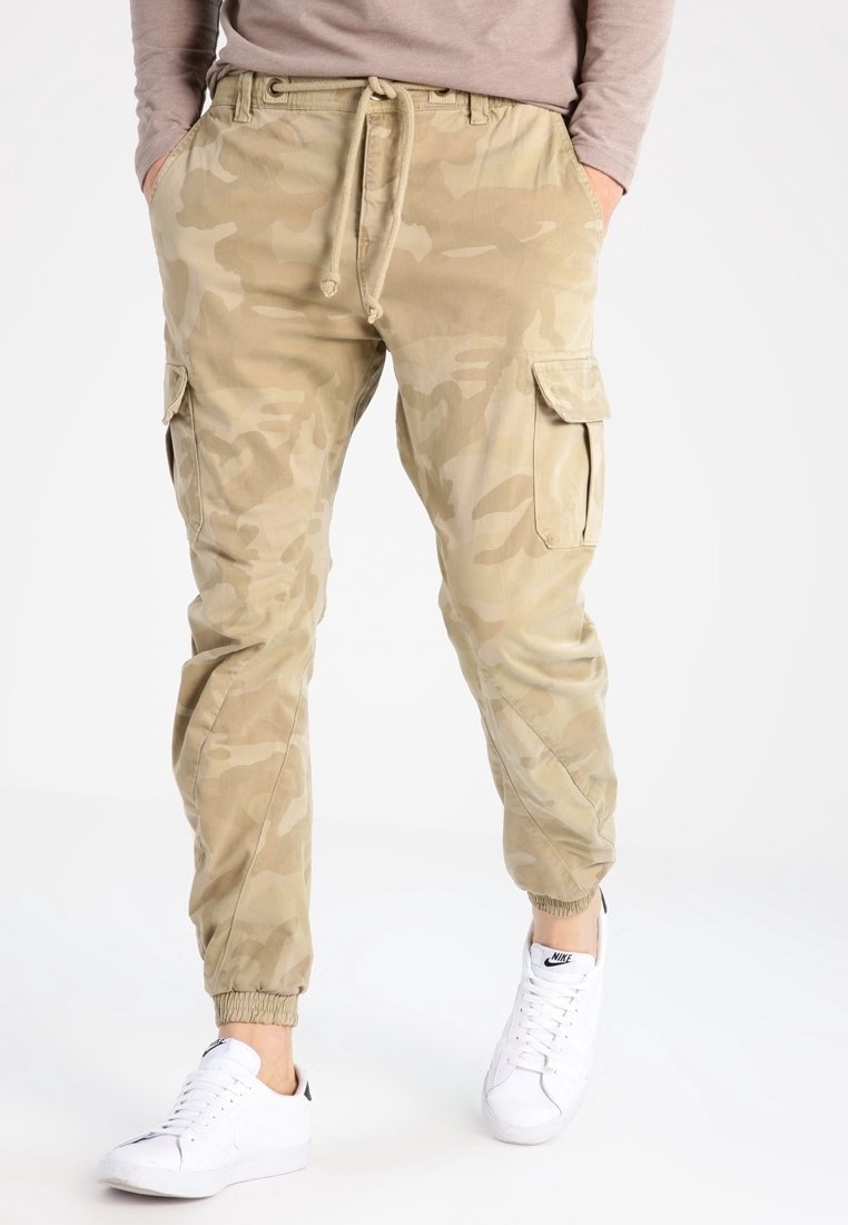 Personne portant un pantalon cargo jogger camouflage beige avec des poignets élastiques, un haut à manches longues beige, et des baskets blanches Nike, debout devant un fond blanc.