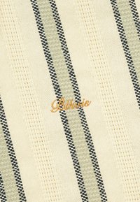 Tissu crème avec des rayures verticales vertes et noires, avec un texte en cursive brodé en or centré sur une rayure.