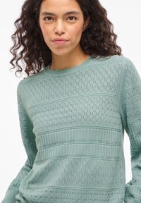 Vrouw met krullend haar, gekleed in een lichtgroene, gestructureerde gebreide trui met ruitpatronen en geribbelde horizontale strepen.