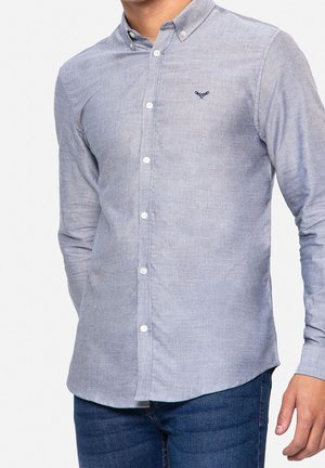 Homme portant une chemise bleu clair boutonnée avec un petit logo sombre brodé sur la poitrine et un jean bleu foncé, debout, les bras détendus.