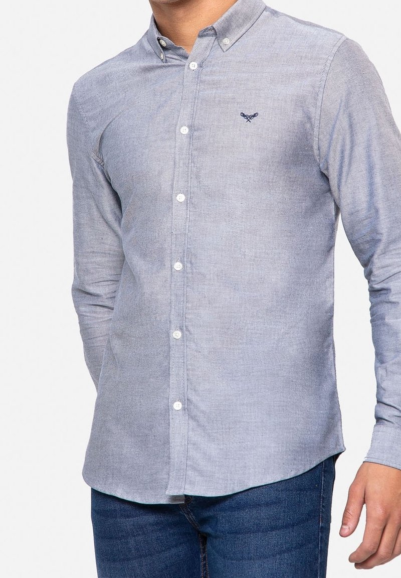 Homme portant une chemise bleu clair boutonnée avec un petit logo sombre brodé sur la poitrine et un jean bleu foncé, debout, les bras détendus.