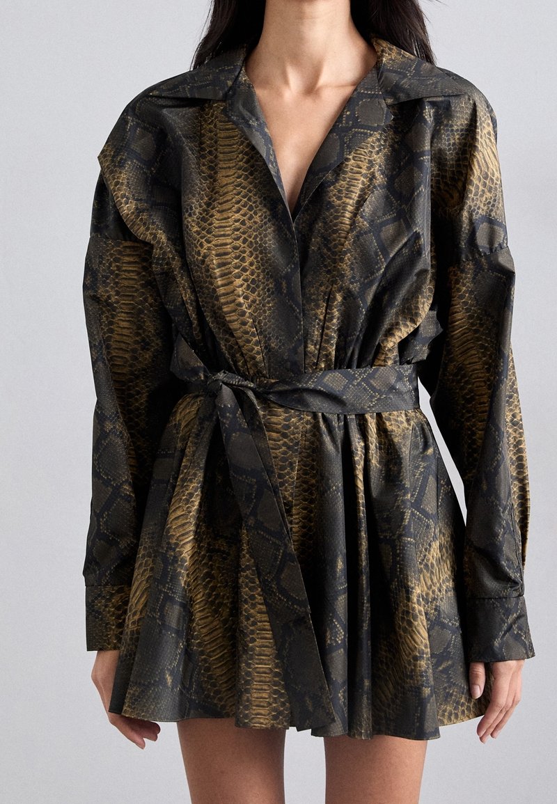 Femme portant une mini-robe à manches longues ceinturée avec un motif en peau de serpent noir et marron, debout devant un fond gris uni.