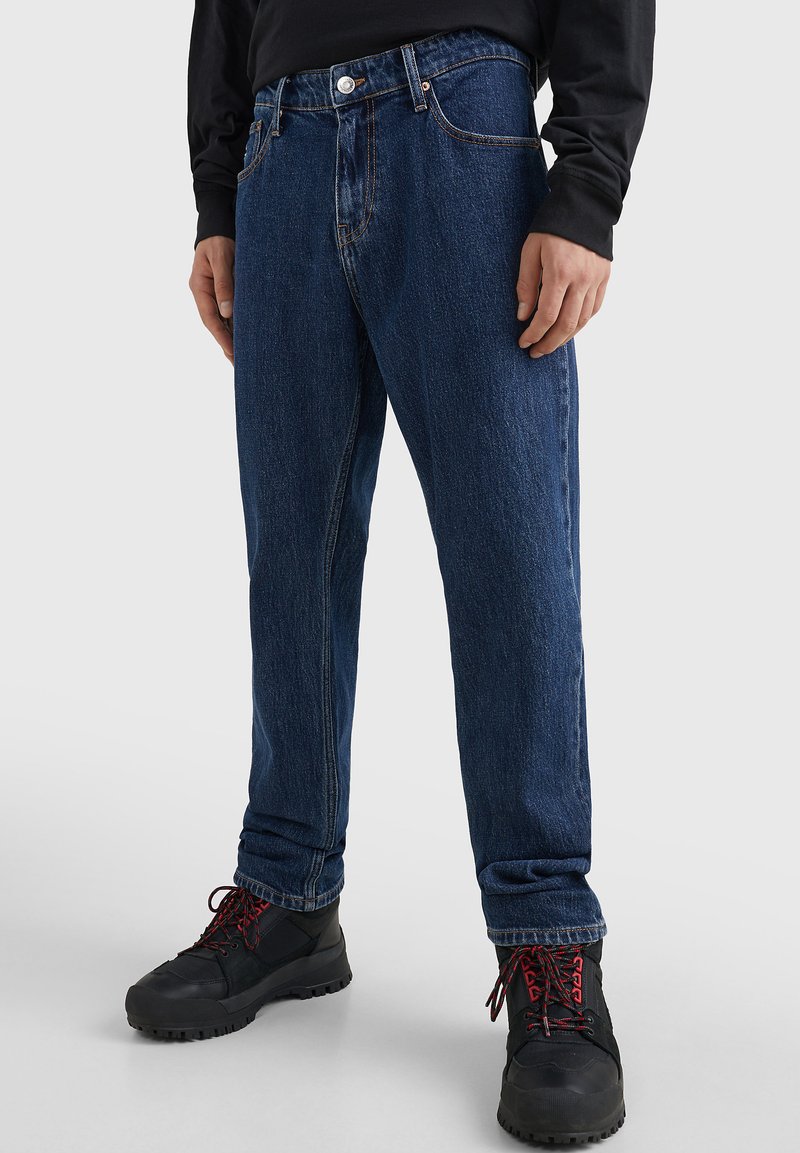 Pantalones vaqueros de mezclilla azul con un corte relajado, que cuentan con un cierre de botones y dos bolsillos frontales. Estilizados con botas negras de caña alta y una camiseta de manga larga negra.