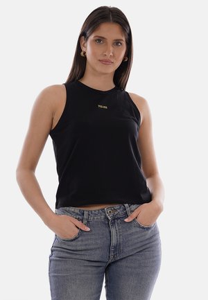 Mujer joven con top sin mangas negro con el logo "YES-ZEE" y jeans azules, de pie con las manos en los bolsillos frente a un fondo liso.