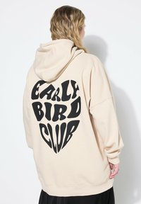 Sudadera beige con un gran gráfico negro en forma de corazón que presenta el texto "EARLY BIRD CLUB" en una tipografía audaz y divertida. Tela suave, corte relajado.
