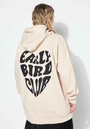 Felpa beige con un grande grafico a forma di cuore nero che presenta il testo "EARLY BIRD CLUB" in un carattere audace e giocoso. Tessuto morbido, vestibilità rilassata.