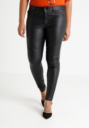 Vrouw die zwarte slim-fit broek van nepleer draagt met geribbelde details op de knieën en zwarte pumps met puntige neus, staand op een witte vloer.
