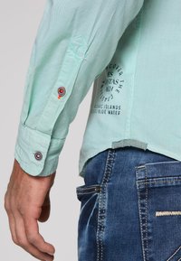 Camicia a maniche lunghe di colore verde chiaro con texture a coste; presenta accenti di bottoni decorativi e dettagli di testo stampati sul lato.
