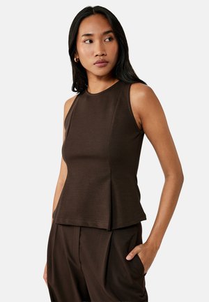 Frau mit langen schwarzen Haaren, die ein ärmelloses dunkelbraunes Top und passende Hosen trägt, steht mit einer Hand in der Tasche und schaut zur Seite.