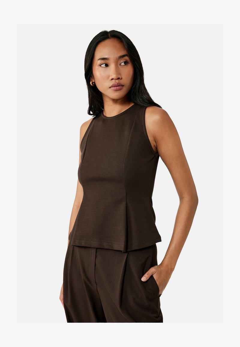 Woman with long black hair wearing a sleeveless dark brown top and matching pants, standing with one hand in pocket, looking to the side.