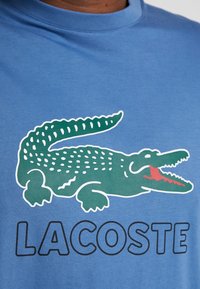 Niebieska bawełniana koszulka z zielonym nadrukiem krokodyla z białym obrysem i czerwonym otworem ustnym oraz napisem "LACOSTE" wyświetlonym na czarno poniżej.