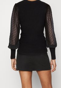 Maglione nero a coste con maniche a sbuffo trasparenti a pois e polsini aderenti, abbinato a una corta gonna nera. In posa da dietro.