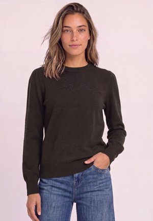 Jeune femme aux cheveux ondulés mi-longs porte un pull foncé à manches longues et un jean bleu, debout avec une main dans sa poche.