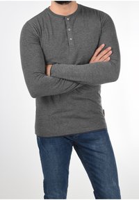 Man in een strak grijs lange mouwen henley-shirt en blauwe jeans met de armen gekruist voor een effen witte achtergrond.