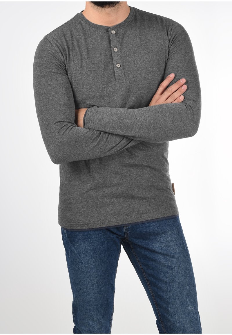 Man in een strak grijs lange mouwen henley-shirt en blauwe jeans met de armen gekruist voor een effen witte achtergrond.